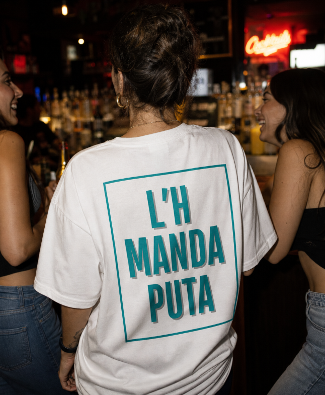 Chica de espaldas en un bar con amigas llevando la camiseta blanca L'H MANDA PUTA